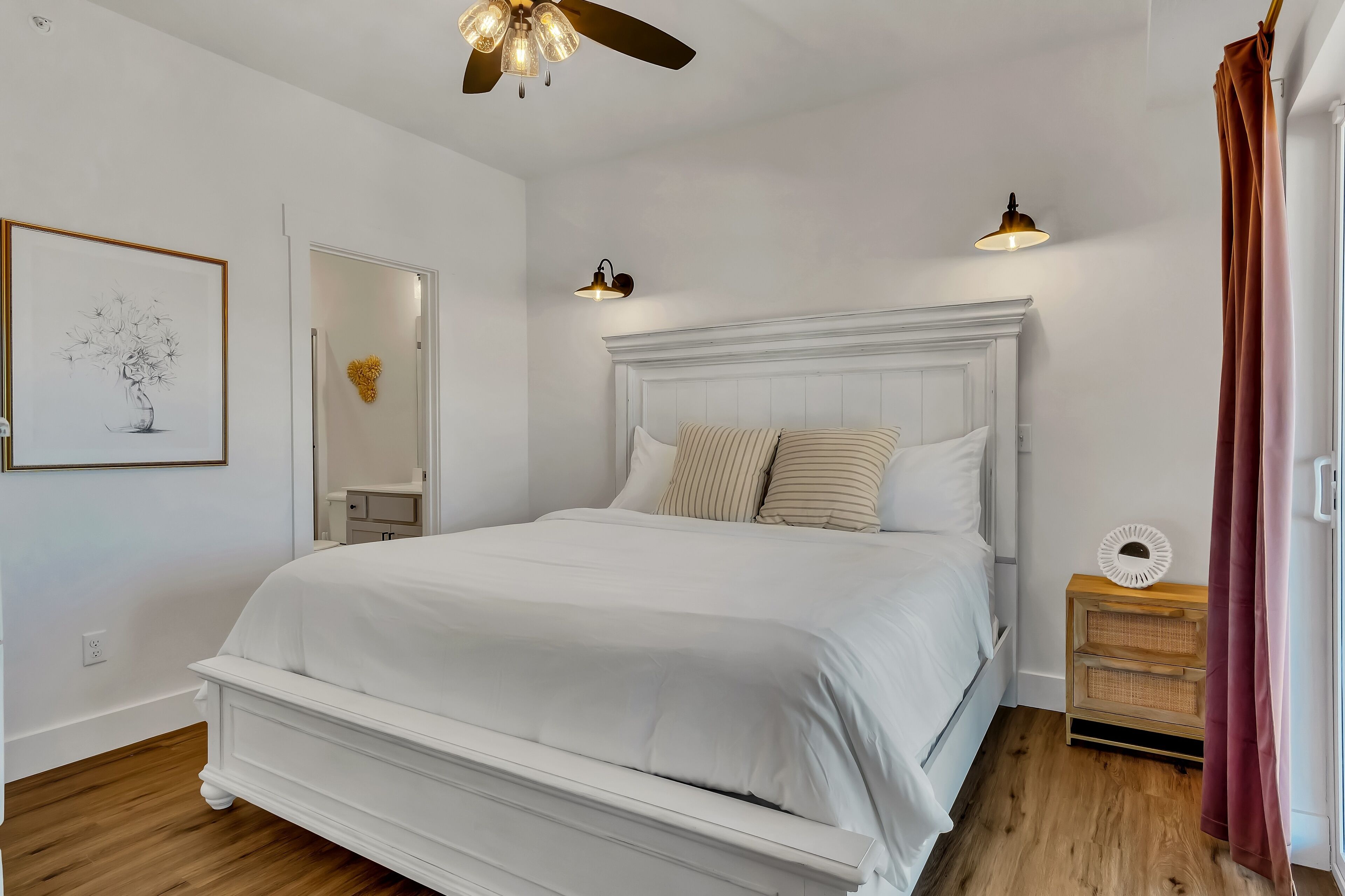 A bedroom with a white bed, a ceiling fan, a bedside table with a lamp, and a framed picture on the wall.