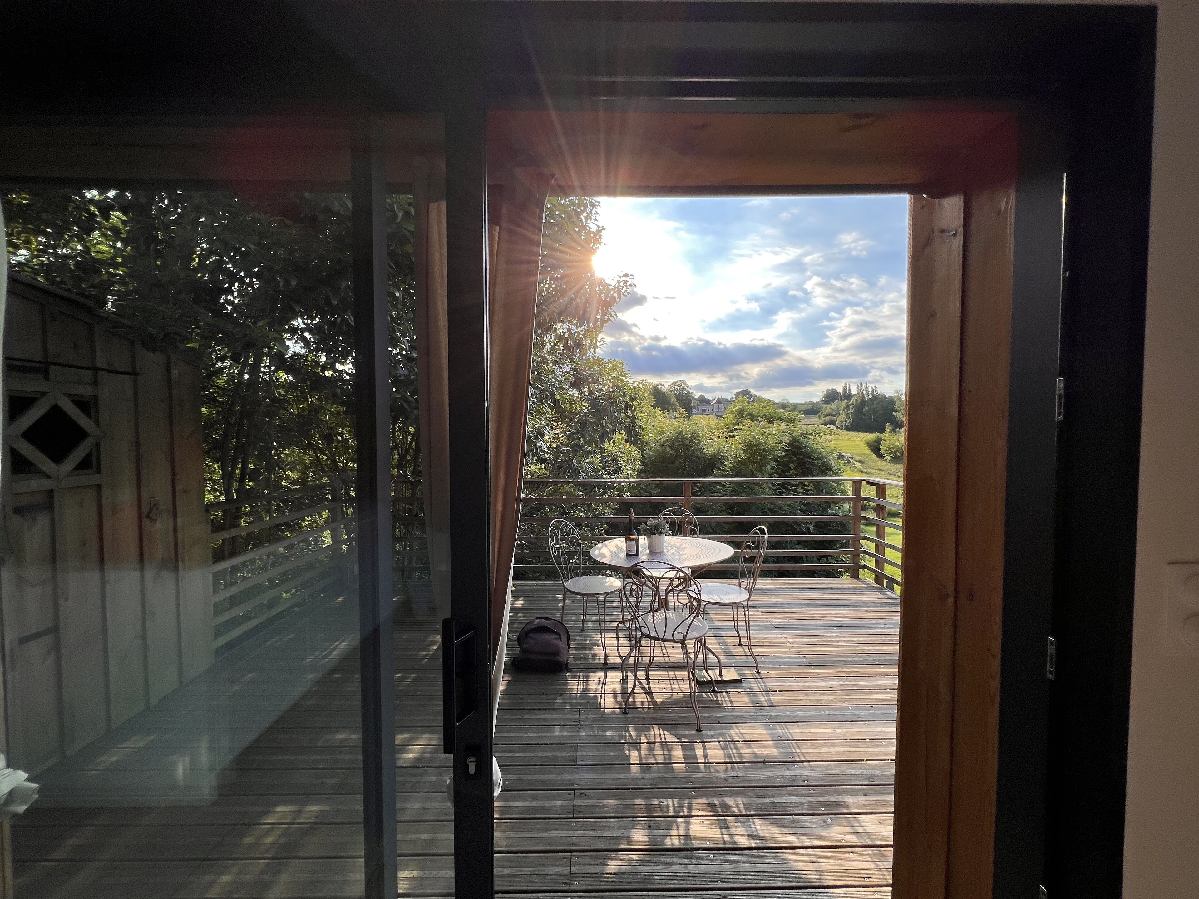 A wooden deck with a table and chairs, a glass door leading to a view of a house and greenery.