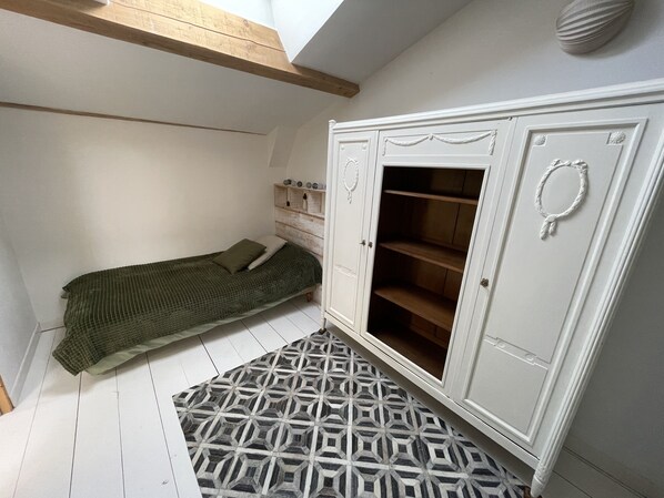 Ein kleines, aufgeräumtes Schlafzimmer mit einem Einzelbett, einem Kleiderschrank mit offenen Regalen und einem gemusterten Boden.