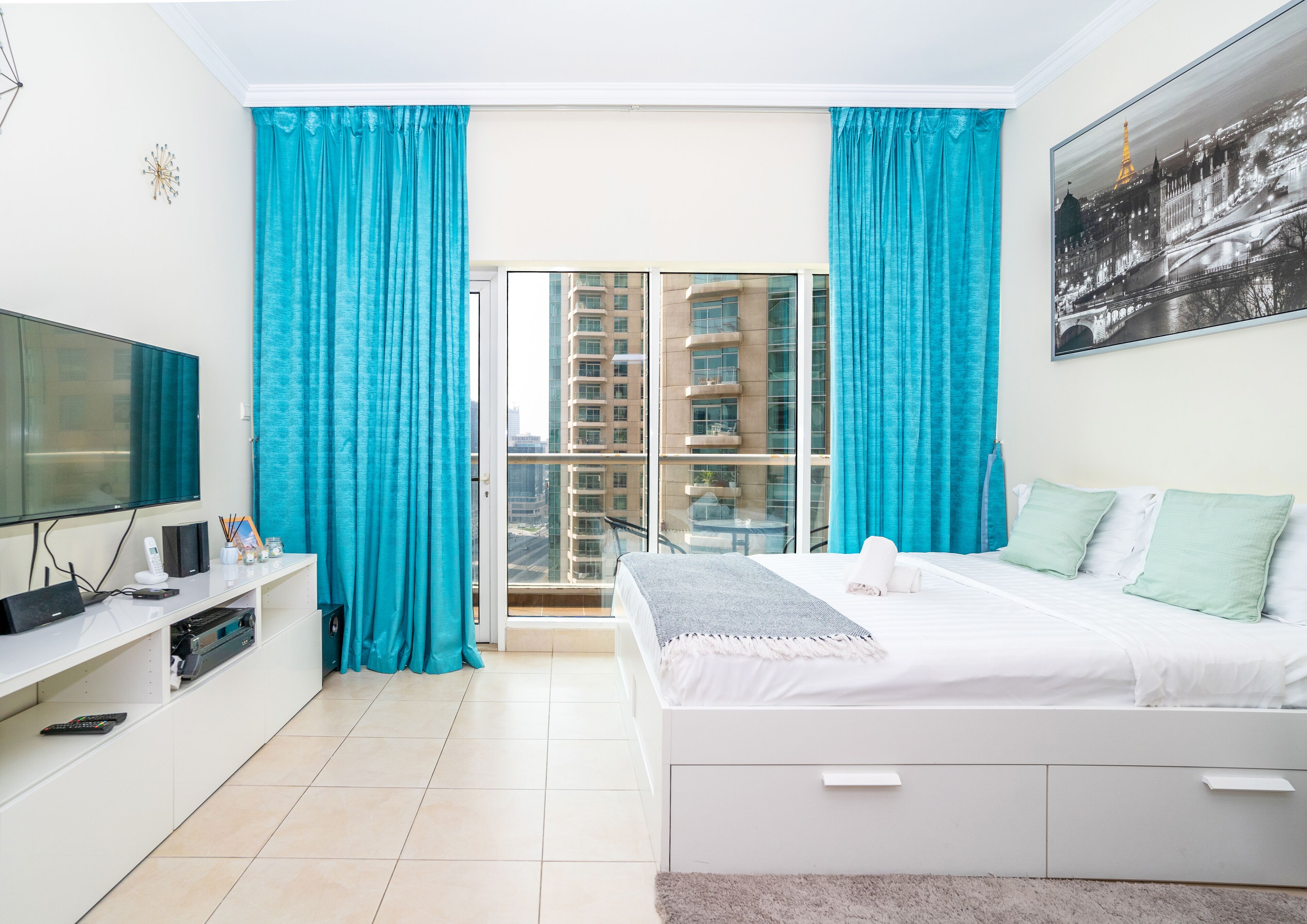 A modern hotel room with a large bed, a TV, a desk with a computer, and a balcony with a city view.