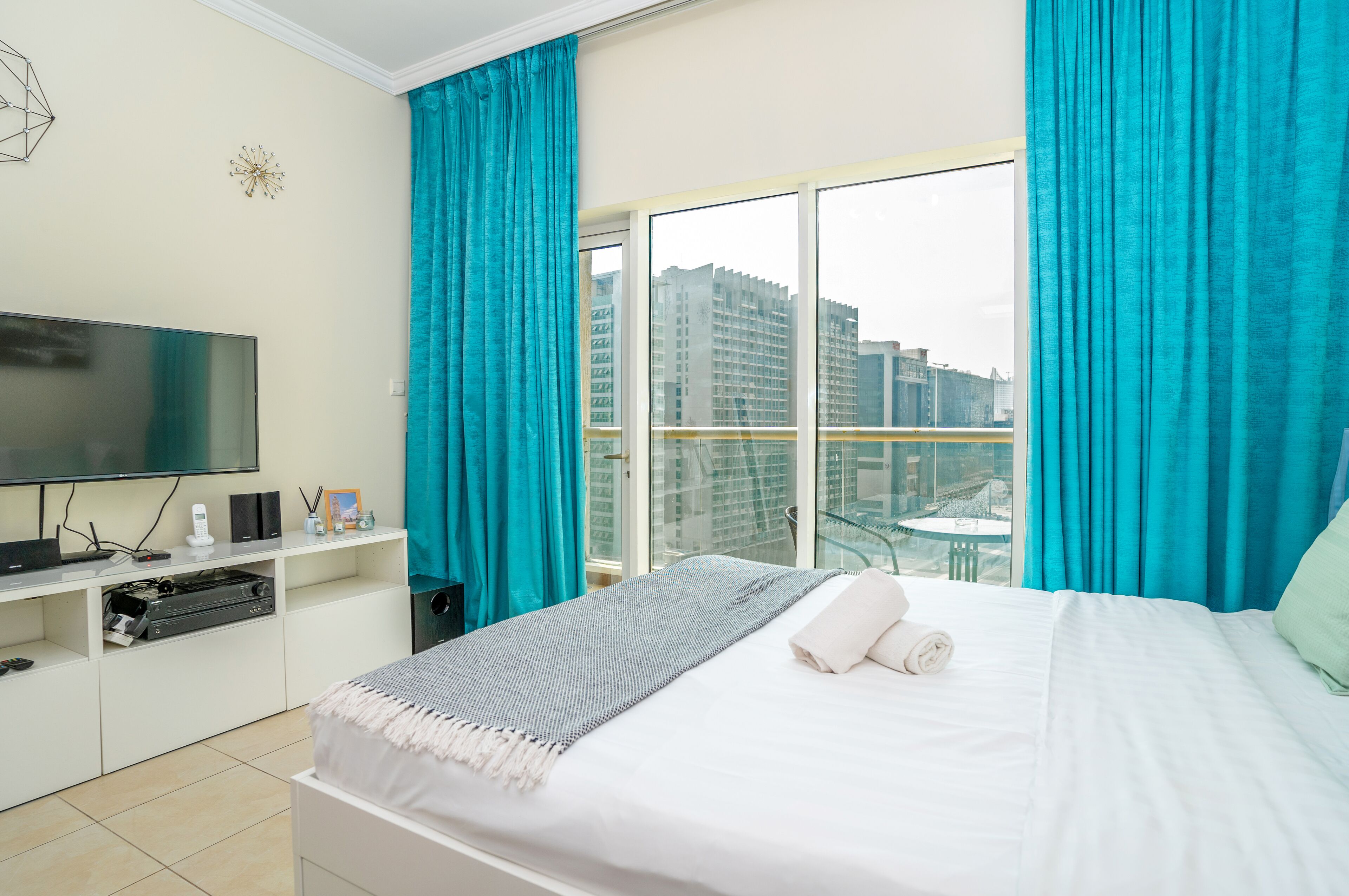 A hotel room with a bed, a TV, a desk with speakers, and a view of buildings through large windows.
