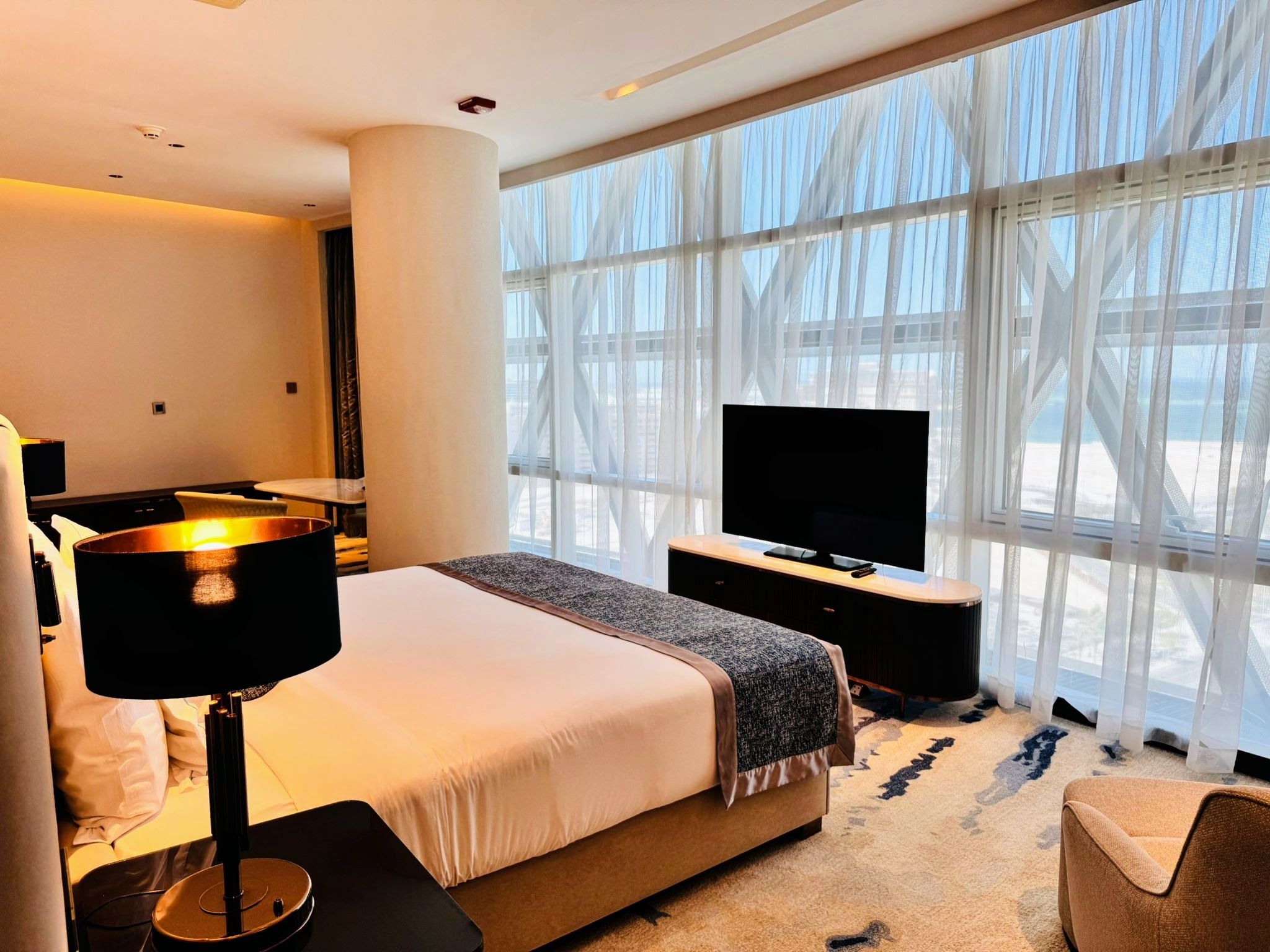 Een moderne hotelkamer met een groot bed, een flatscreen televisie, een bureau en een groot raam met dunne gordijnen.