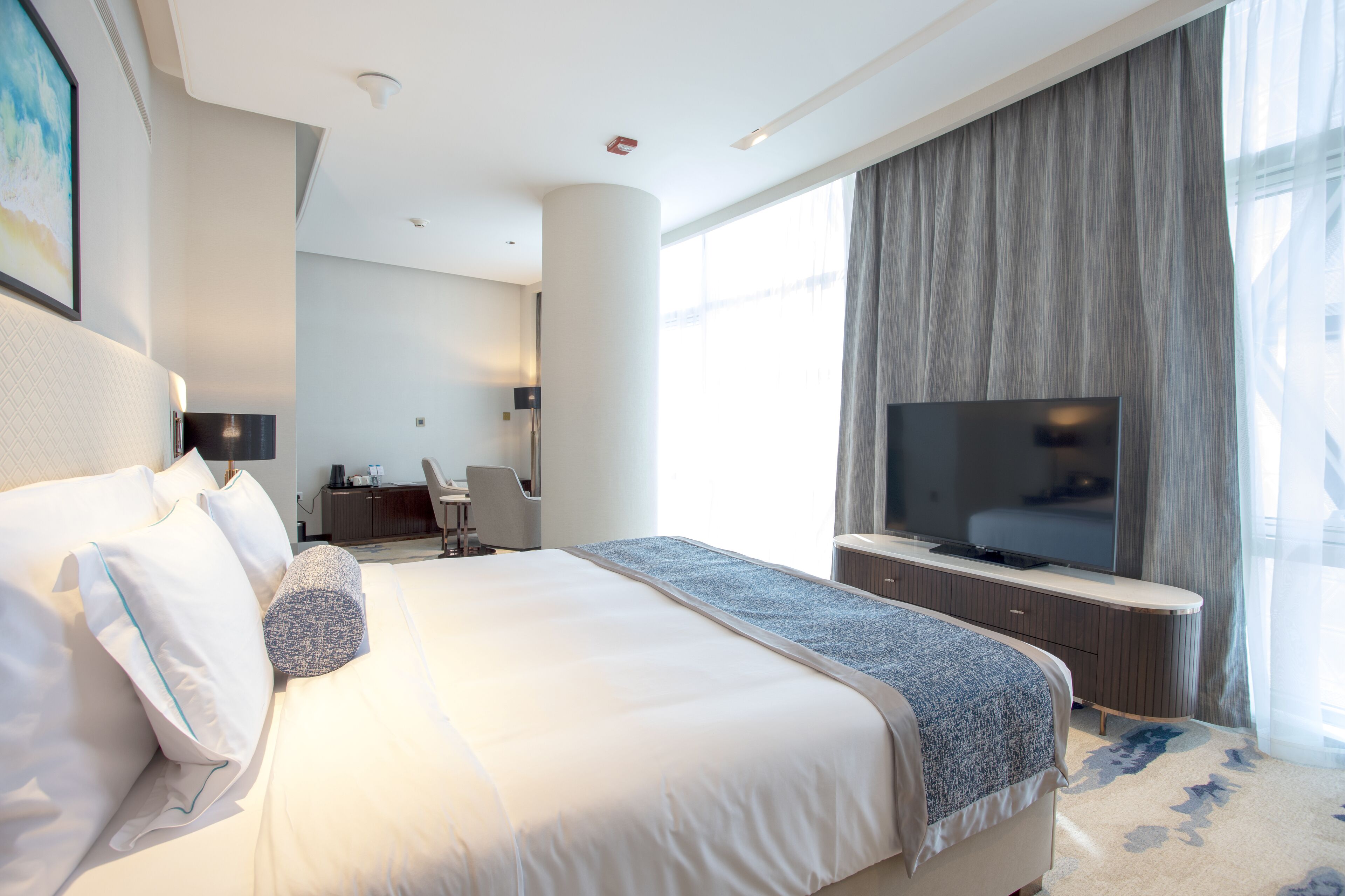 Quarto de hotel moderno com uma cama grande, televisão de tela plana, escrivaninha e uma janela ampla com cortinas.