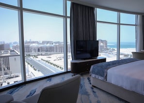 Habitación de hotel con un ventanal grande, una cama, un televisor y vistas a la ciudad y a la playa.