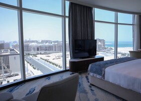 Hotelkamer met groot raam, bed, televisie en uitzicht op stadsbeeld en strand.
