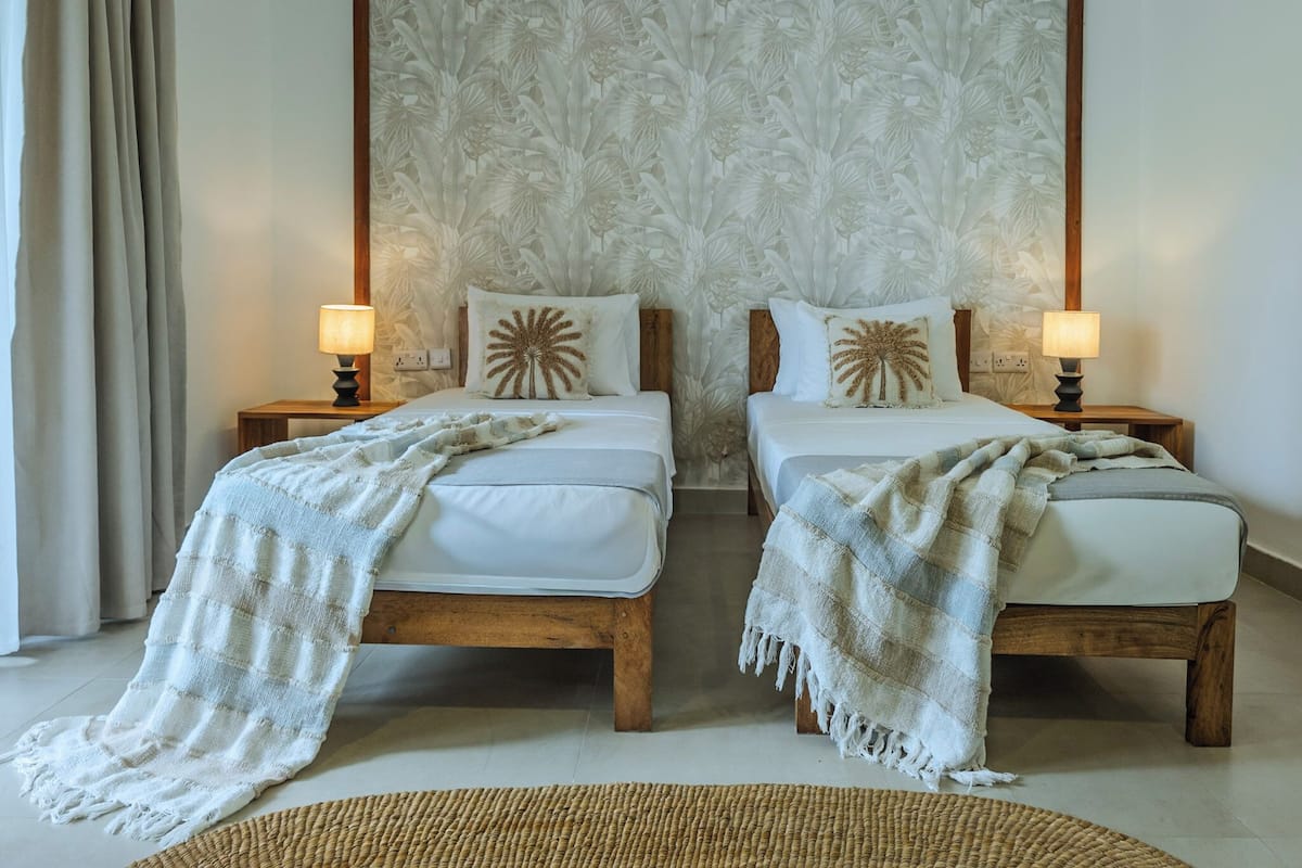 Una habitación con dos camas, un cabecero de gran tamaño con estampado floral, lámparas de noche y una alfombra tejida.