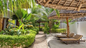 A tropical garden with a thatched roof structure, a gravel path, and lounge chairs. - Tamu Tamu Boutique Hotel (Pwani Mchangani)