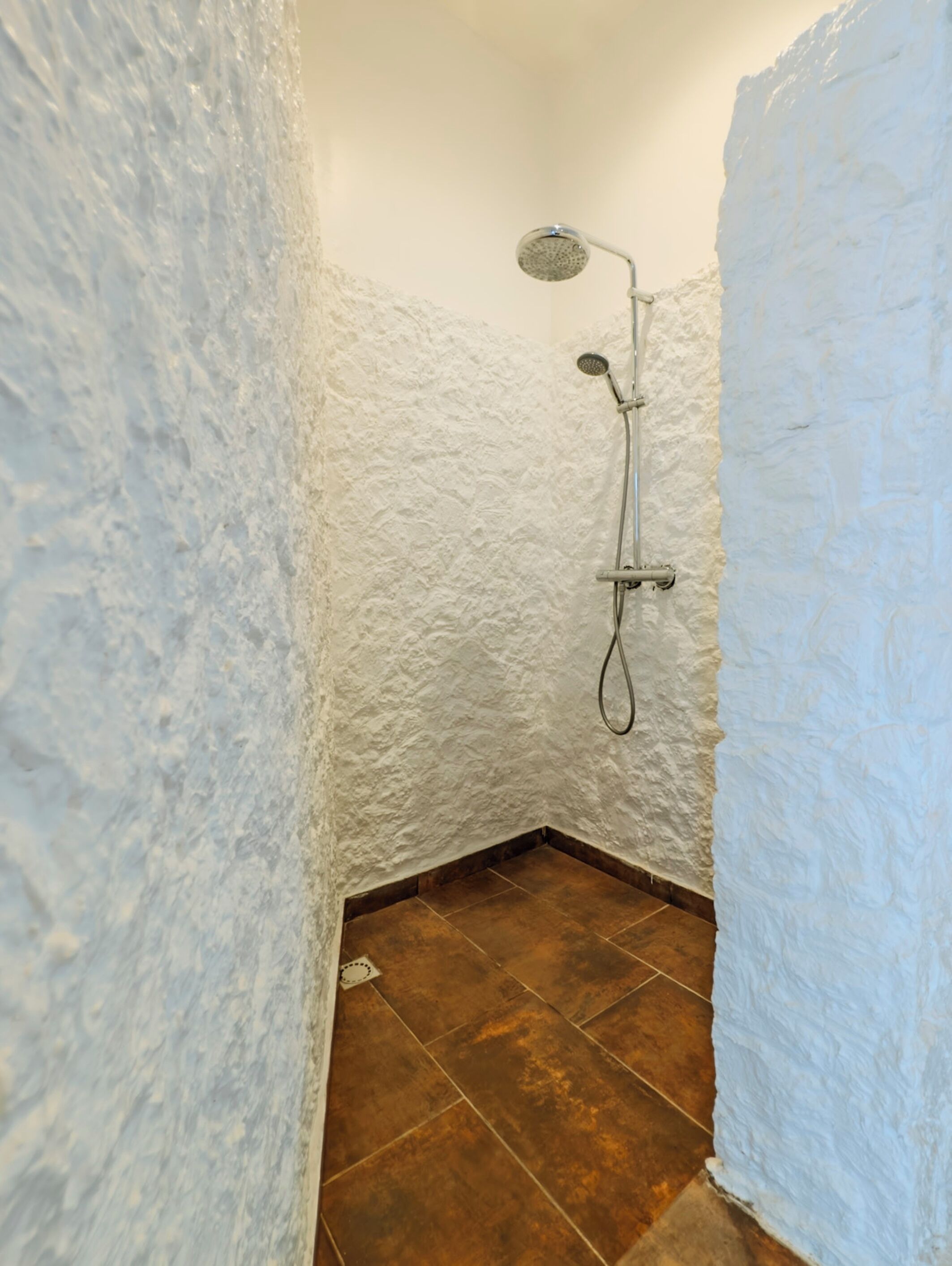 Une douche d’angle avec des murs blancs à texture et un sol carrelé marron.