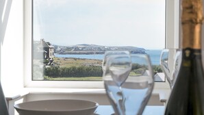 Ett matbord med ett glas vatten och en flaska champagne, ett fönster med utsikt över kusten och en stol.
