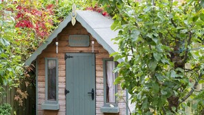 Eine kleine Holzhütte mit grüner Tür und zwei Fenstern, umgeben von üppigem Grün.