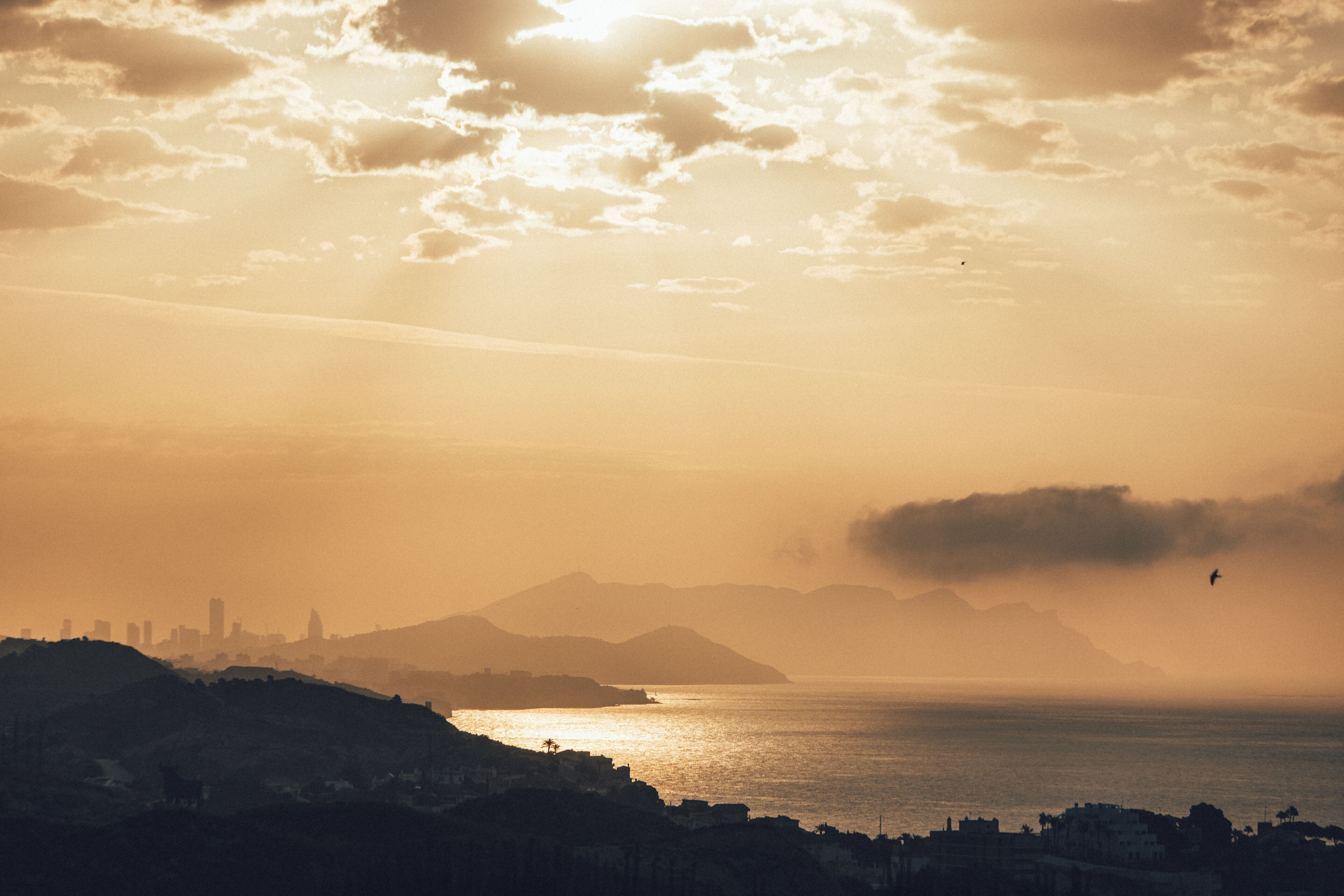 A coastal cityscape at sunset with a view of the ocean and distant mountains.