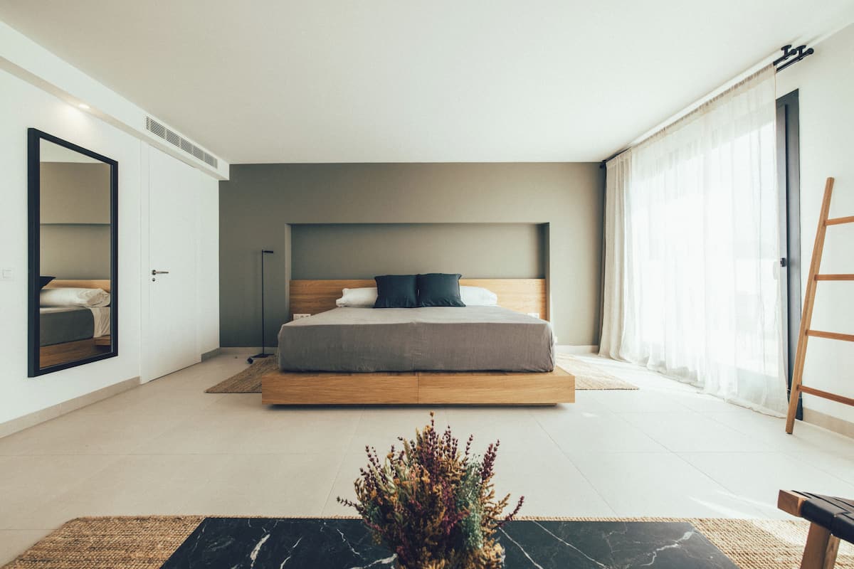 Ein modernes Schlafzimmer mit einem großen Bett, einem Spiegel, einer Stehlampe und Blick ins Freie durch ein Fenster.