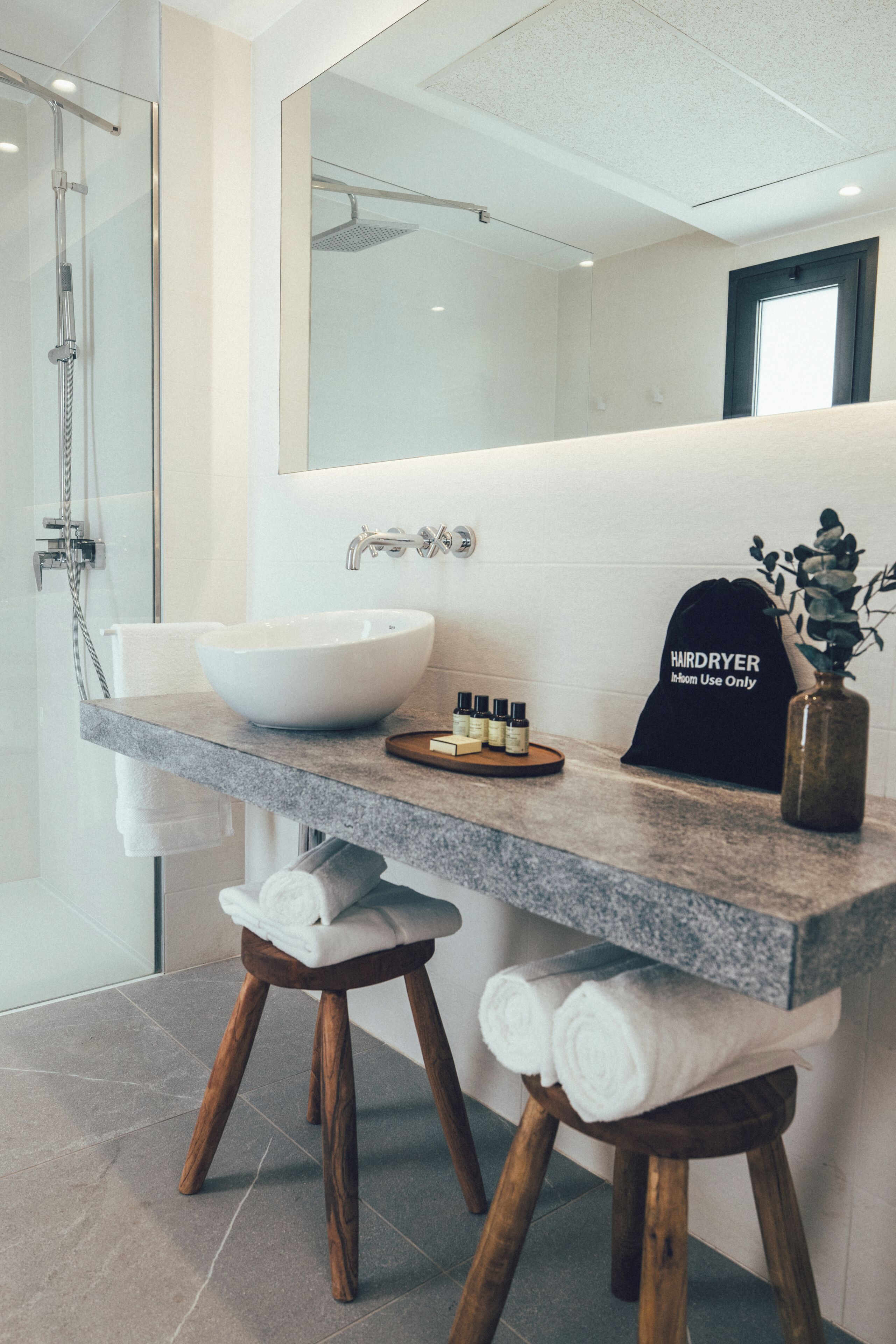 Un baño moderno con un espejo grande, un lavabo sobre encimera y una ducha. Hay toallas sobre taburetes y un secador de pelo en la encimera.