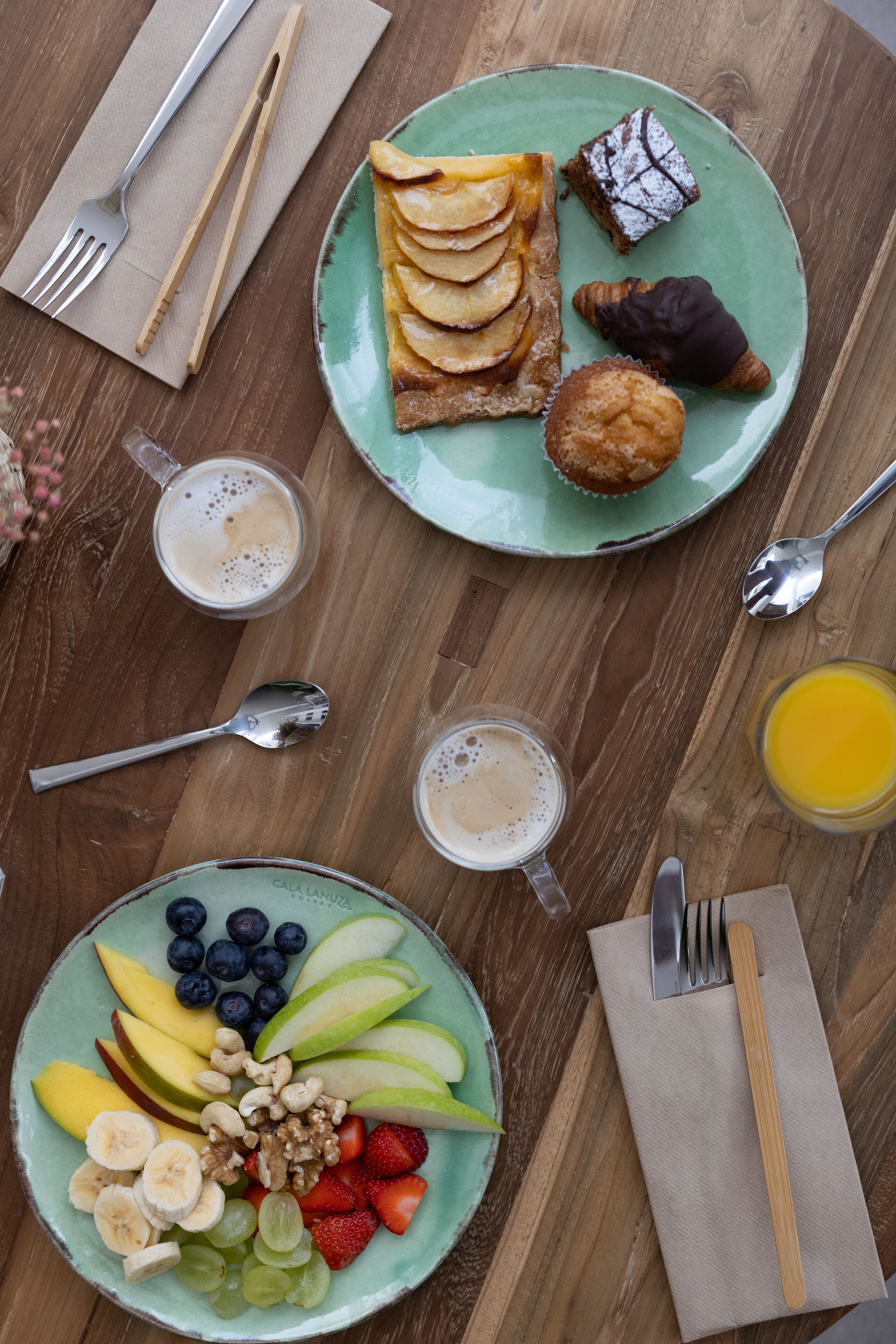 Un desayuno con platos de fruta, pasteles y bebidas sobre una mesa de madera.