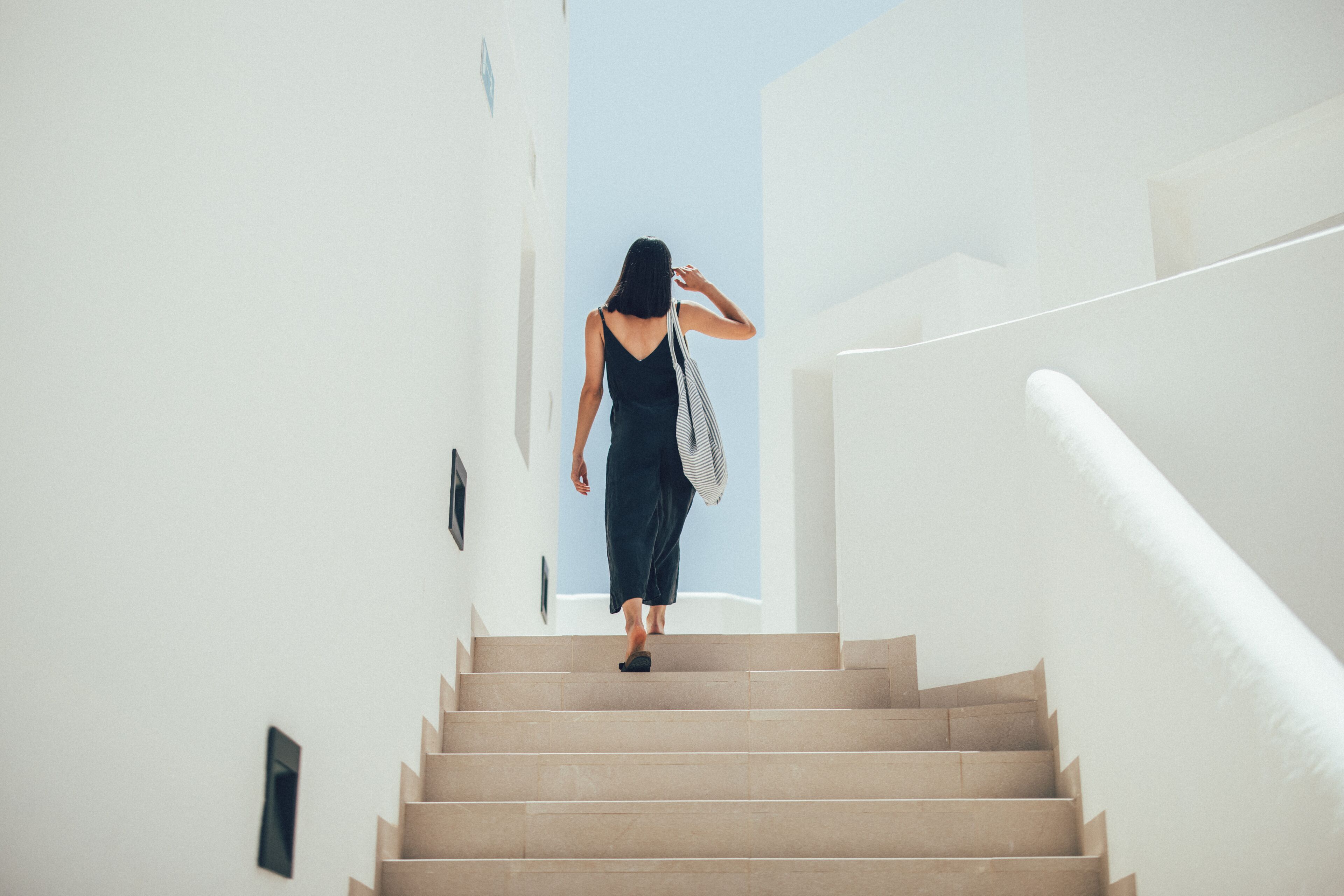 Une femme qui monte un escalier dans un bâtiment aux murs blancs, avec un ciel bleu clair en arrière-plan.