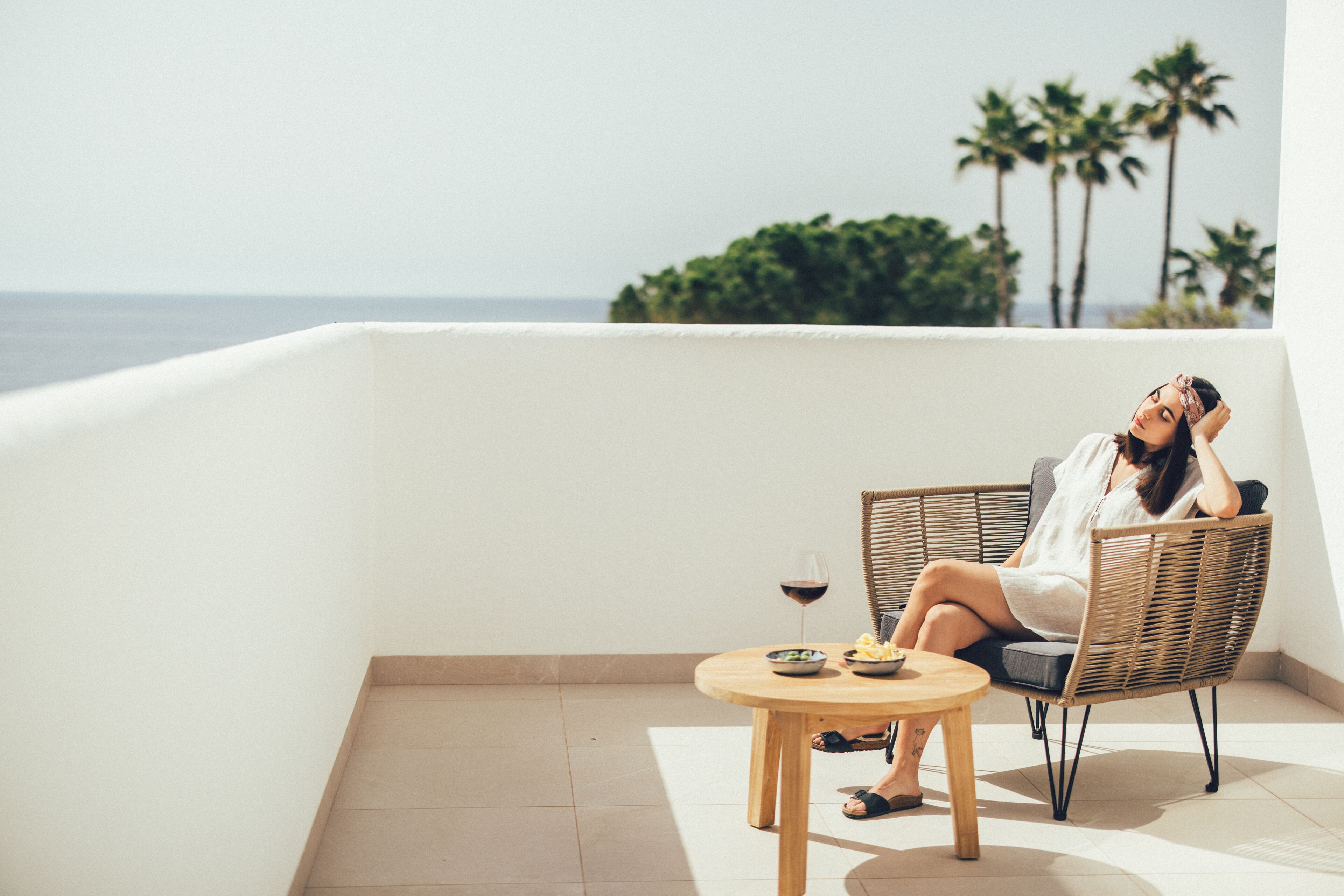 Una donna seduta su una sedia in vimini su un balcone con un tavolo e una ciotola di cibo, con vista su una spiaggia con palme.