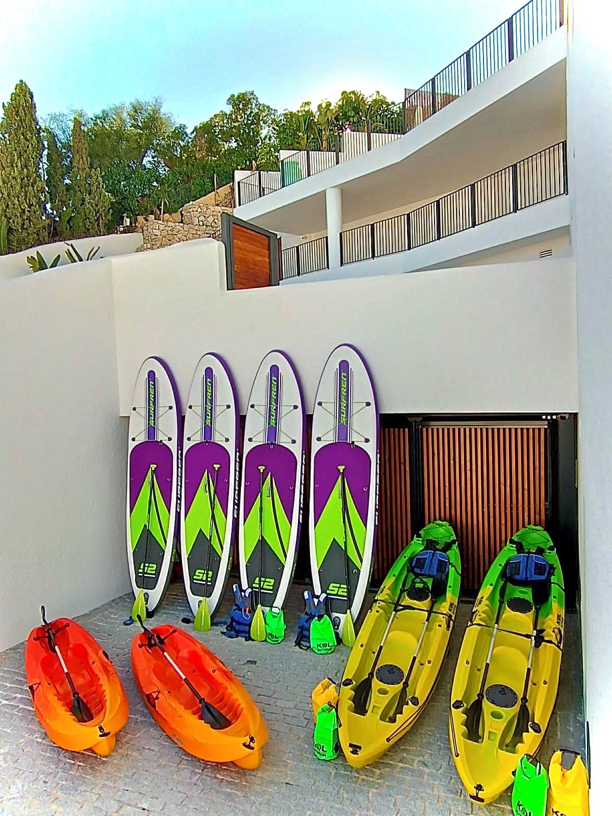 Kayaks y tablas de remo alineadas frente a un edificio con pared de piedra y balcón.