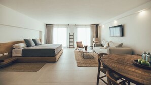 A modern hotel room with a large bed, a sofa, a dining table, and a TV. - Cala Lanuza Suites (El Campello)