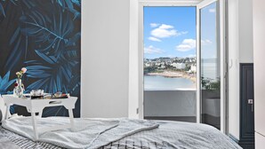 Een slaapkamer met een bed, een wit bureau, een vaas met bloemen en uitzicht op een strand en gebouwen door een geopende deur.