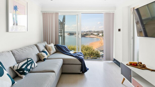 Een moderne woonkamer met een bank, een televisie en uitzicht op strand en stadsbeeld door grote ramen.