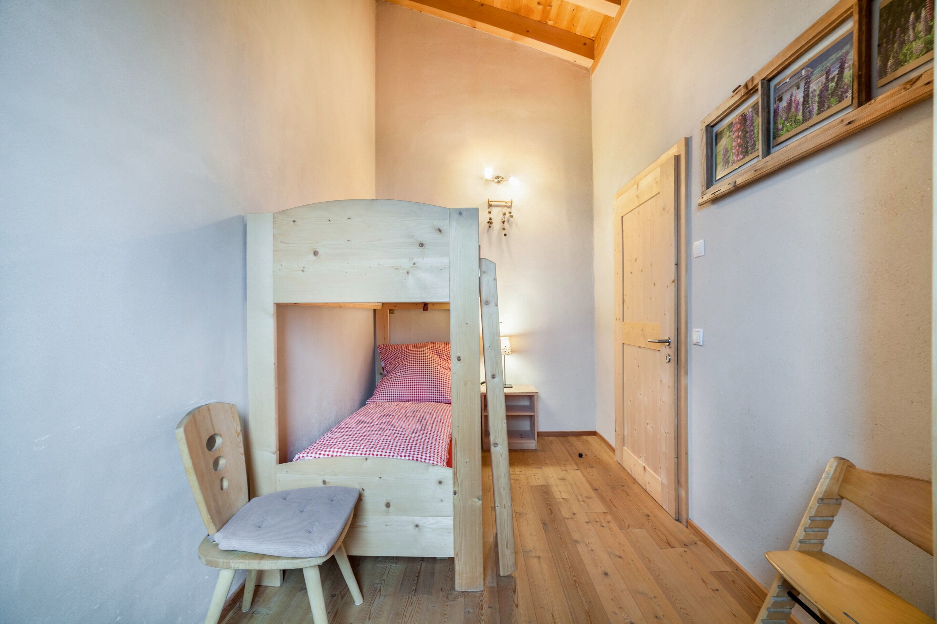 Ein hölzernes Etagenbett mit karierter Bettdecke, ein Holzstuhl mit Kissen und eine hölzerne Bank.