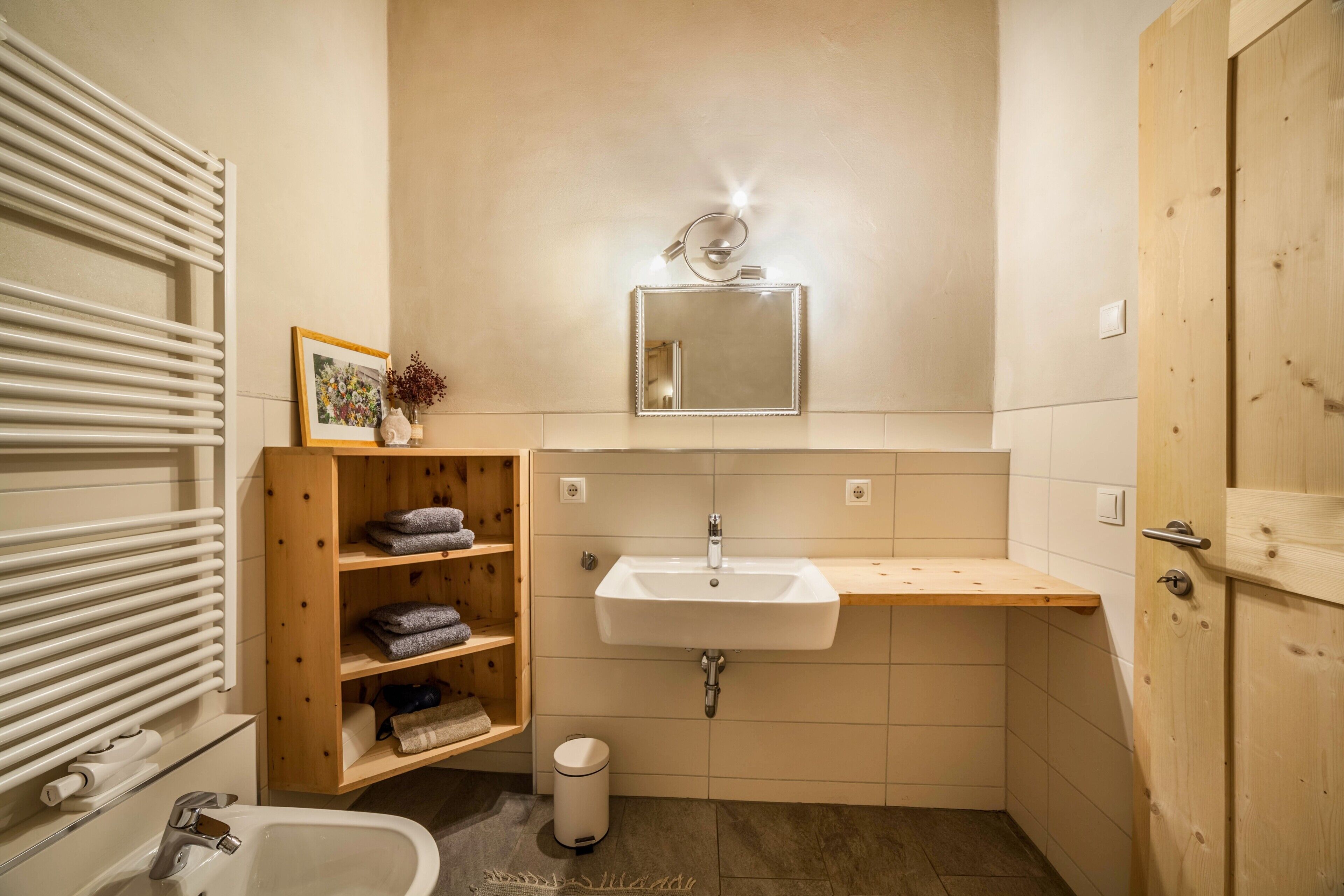 Un baño con lavamanos blanco, repisas de madera, un espejo y un perchero para toallas.