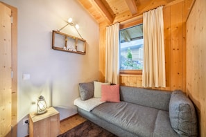 Una habitación con paneles de madera, un sofá gris, una mesita con un farol y una ventana con cortinas.