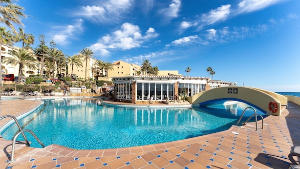 Ein Pool mit gefliestem Sonnendeck, Palmen und einem Gebäude mit großen Fenstern.