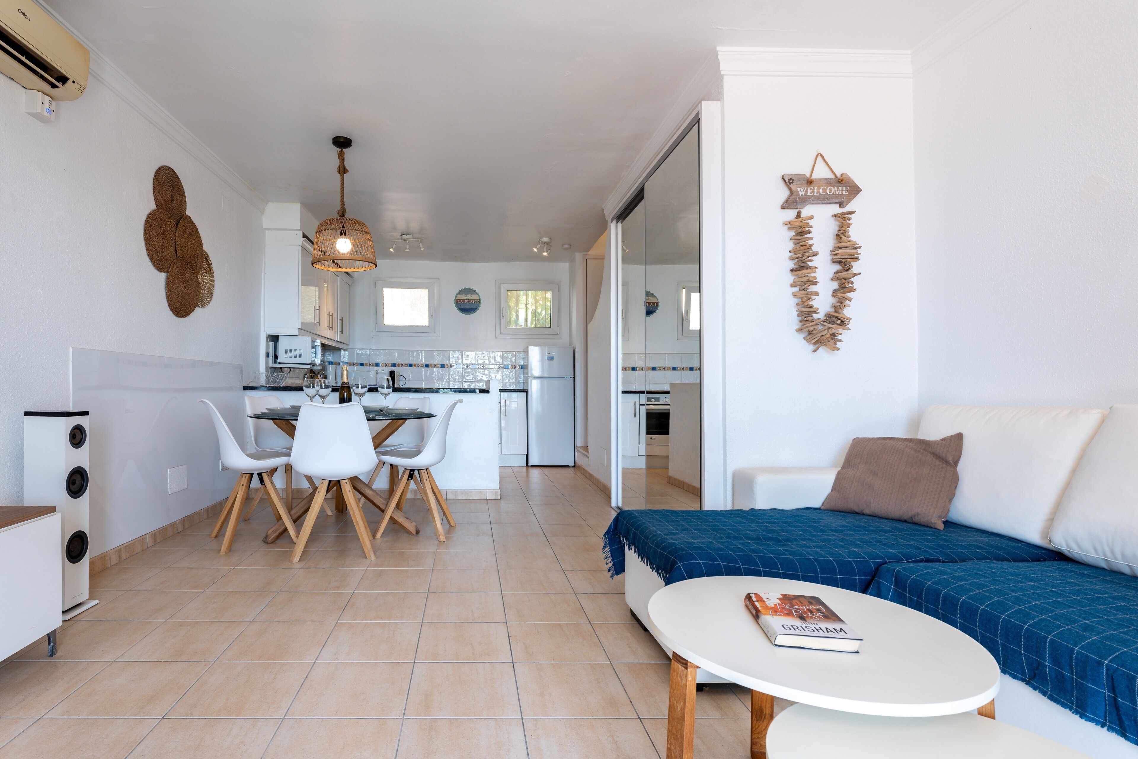 Una sala de estar moderna con un sofá, una mesa de comedor con sillas, una cocina al fondo y un letrero que dice 'Welcome' en la pared.