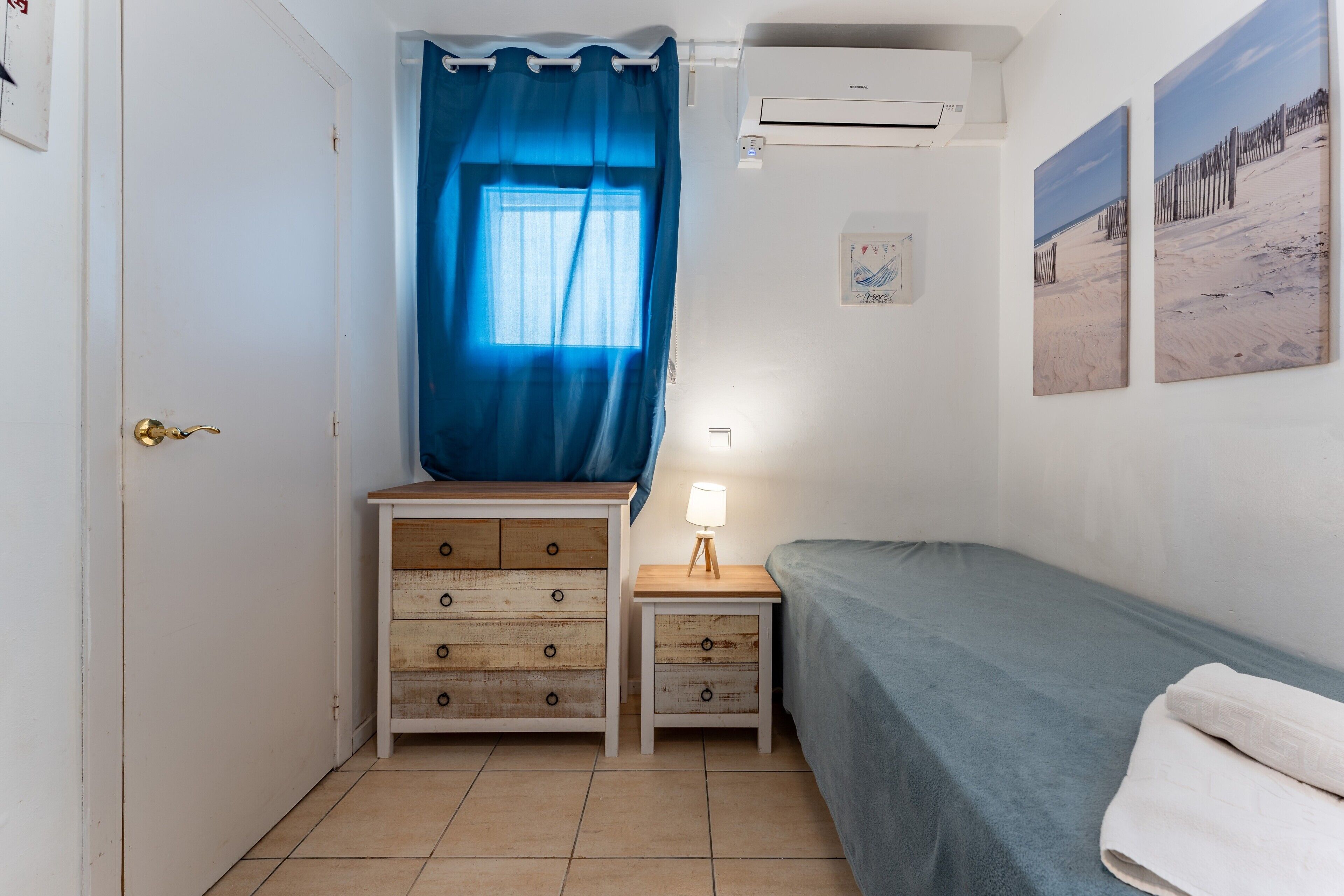 A small, tidy room with a bed, a nightstand, a dresser, a window with blue curtains, and wall art.