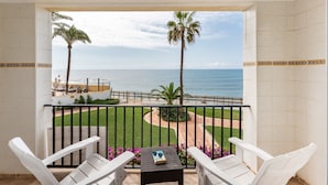 Ein Balkon mit weißen Stühlen, einem kleinen Tisch und Blick auf das Meer und Palmen.