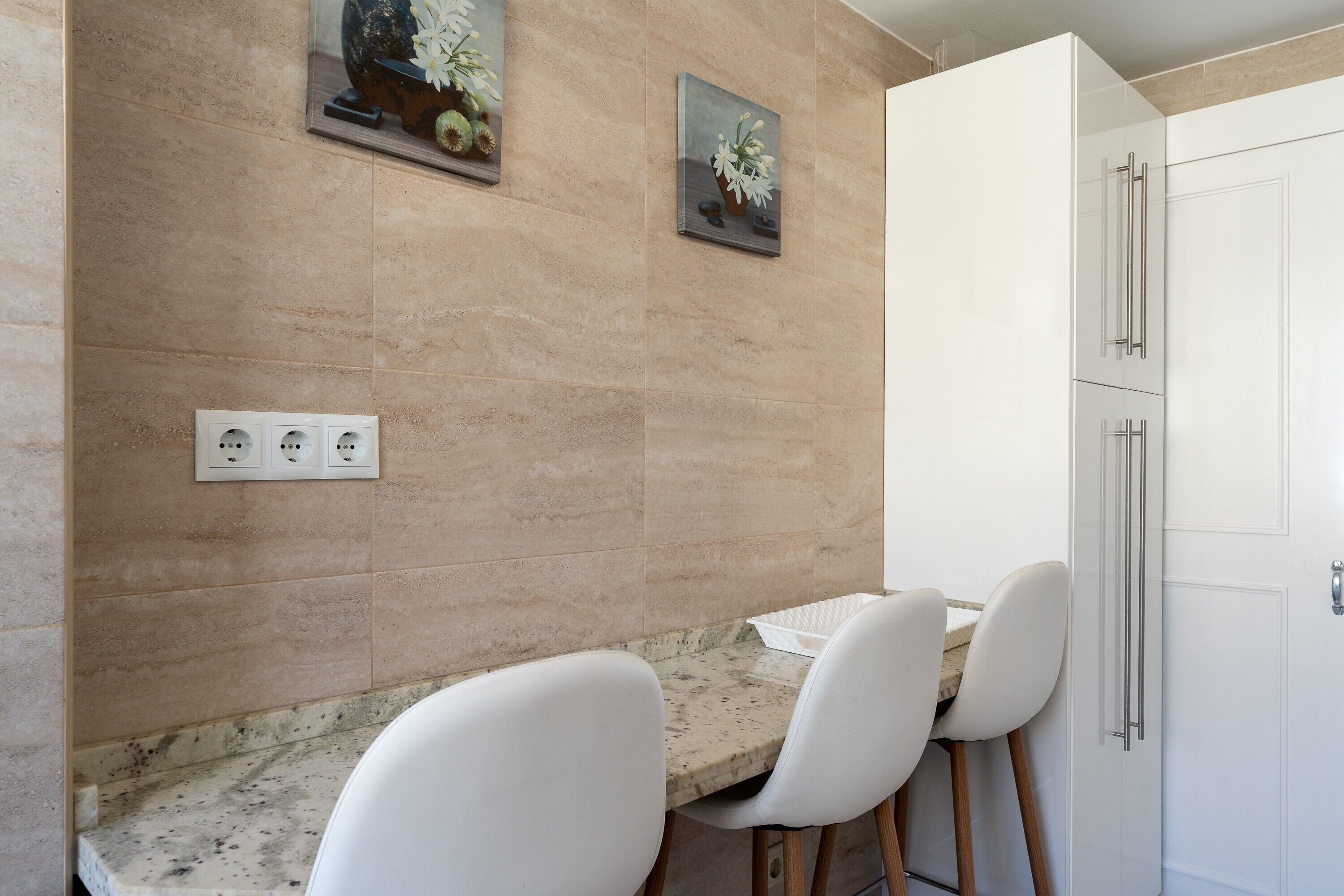Una cocina con encimeras de mármol, sillas blancas, un armario blanco y dos cuadros de flores colgados en la pared.