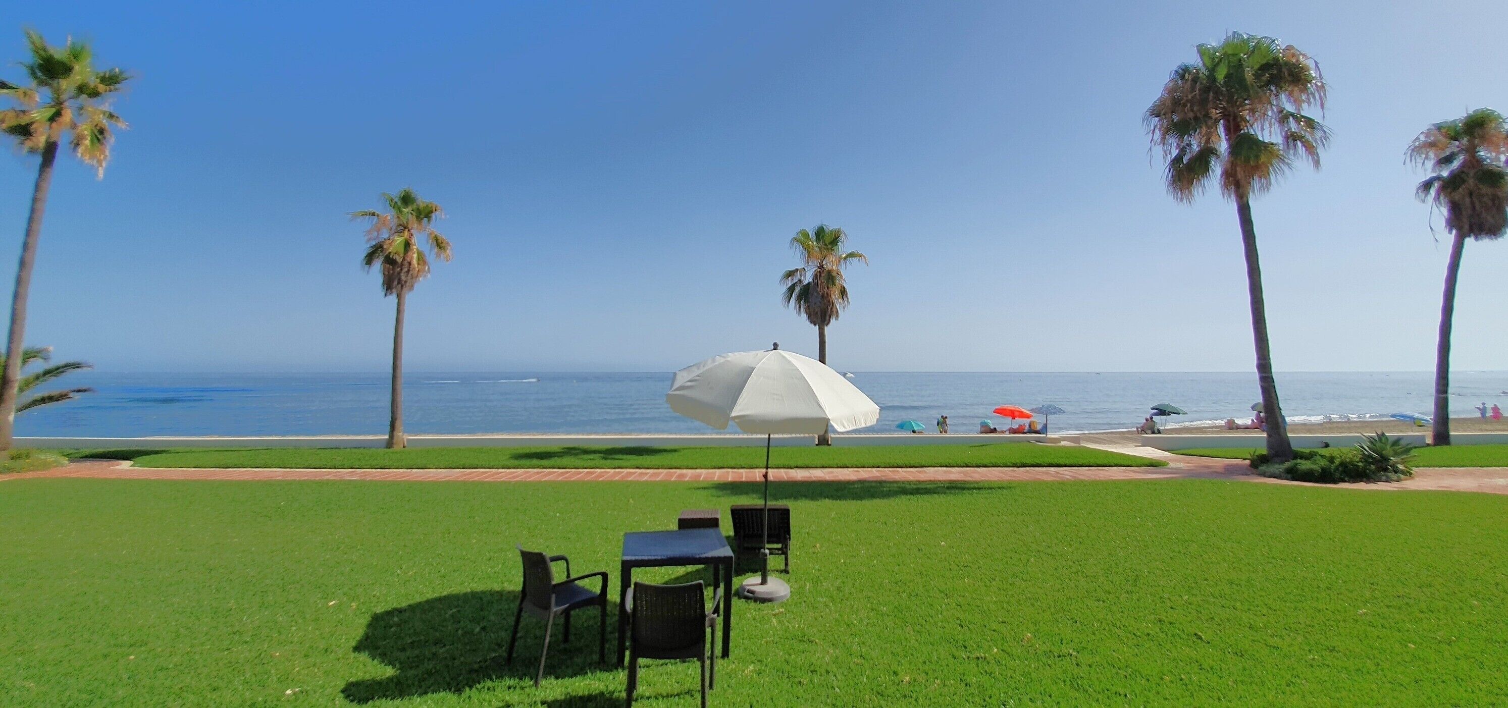 Un espace verdoyant avec un parasol blanc, des chaises et une vue sur l’océan.