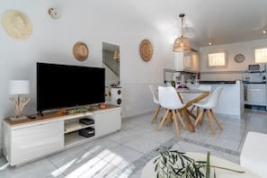 A modern living room with a flat-screen TV, dining area with chairs, kitchen with appliances, and decorative wall items.