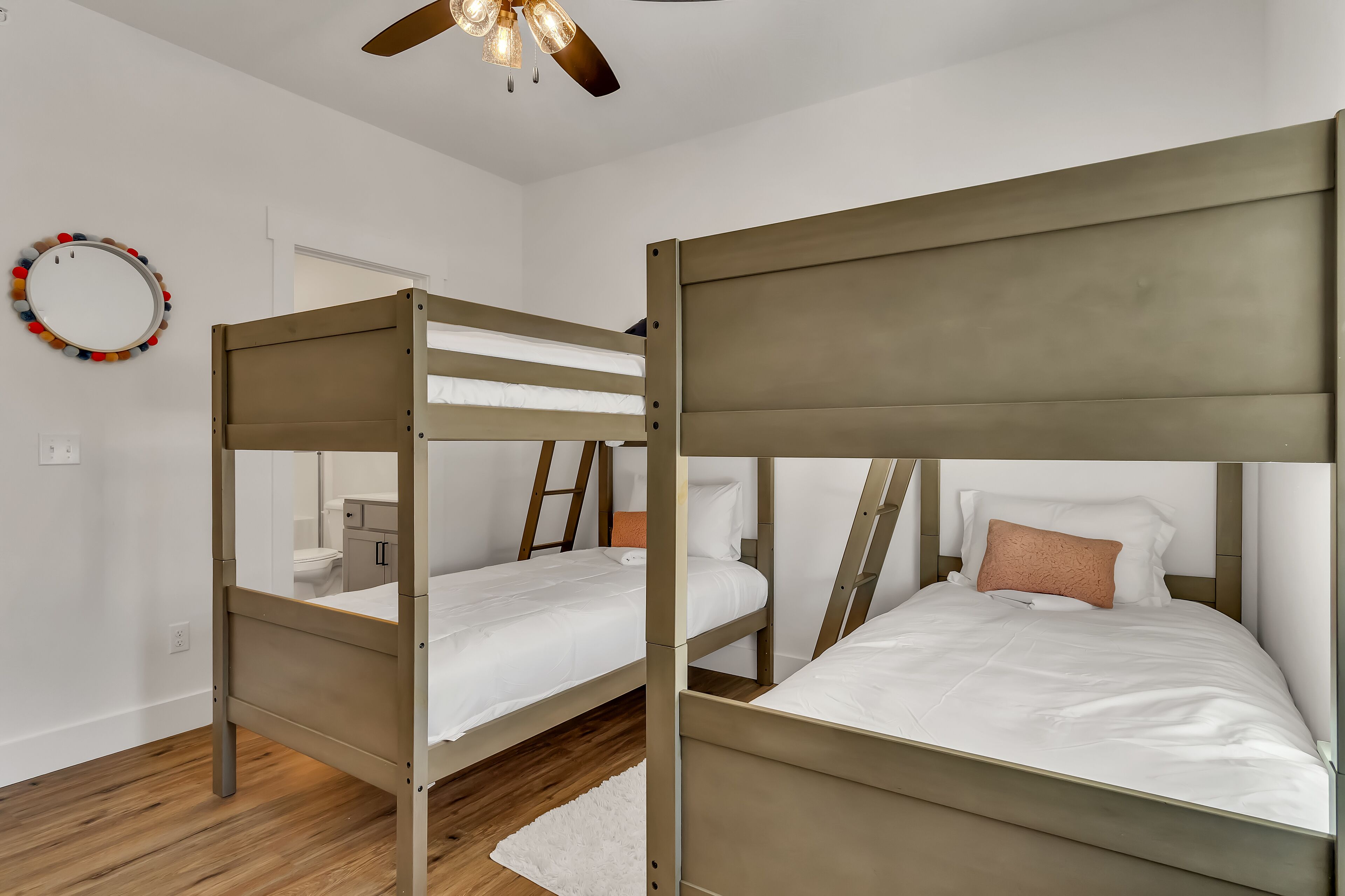 Une chambre avec deux lits superposés, un ventilateur de plafond, une porte blanche et un miroir rond orné de pompons colorés.