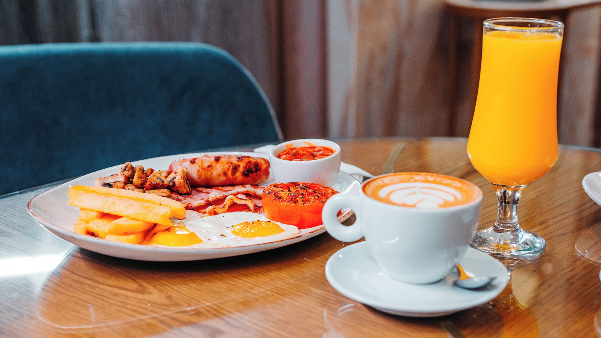 A breakfast plate with eggs, bacon, sausages, and fries, a cup of coffee, and a glass of orange juice on a wooden table.