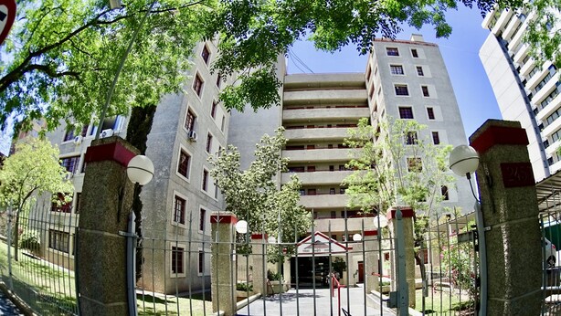 Un immeuble à plusieurs étages doté d’une entrée centrale, entouré d’une clôture en métal et d’arbres.