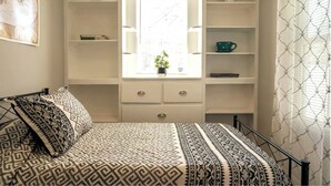 Un dormitorio con cama, ventana con vistas, estante con objetos decorativos y cortina estampada.