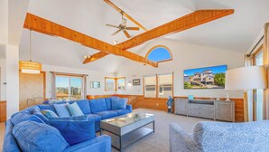 Ein großzügiges Wohnzimmer mit einem blauen Sofa, einem Glastisch, Holzbalken, einem Deckenventilator und einem großen Fenster mit Blick nach draußen.