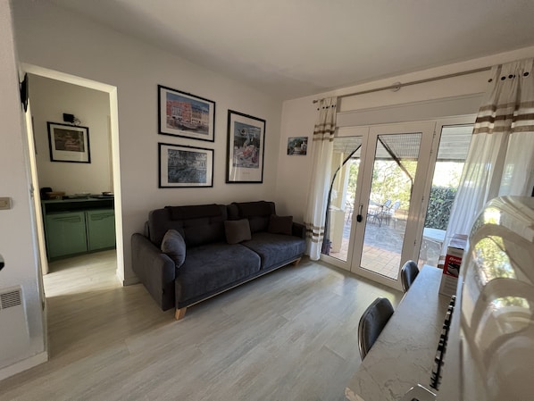 A living room with a grey sofa, a dining table, and a kitchen area visible through an open door.