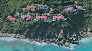 An aerial view of a coastal property with multiple houses, each featuring a red roof, situated on terraced land with a rocky shoreline. - Villa Morning Glory - Private Pool - Amazing Views (St Thomas)
