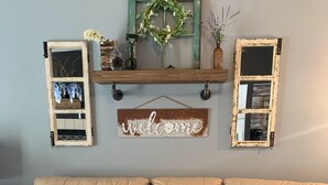 Un sofá de cuero color beige con un cojín a rayas, una cesta de mimbre y decoración rústica, incluyendo un cartel que dice 'welcome' y dos ventanas enmarcadas.