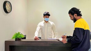 Un comptoir d’accueil avec une horloge au mur, une plante en pot et une personne qui écrit sur un bloc-notes.