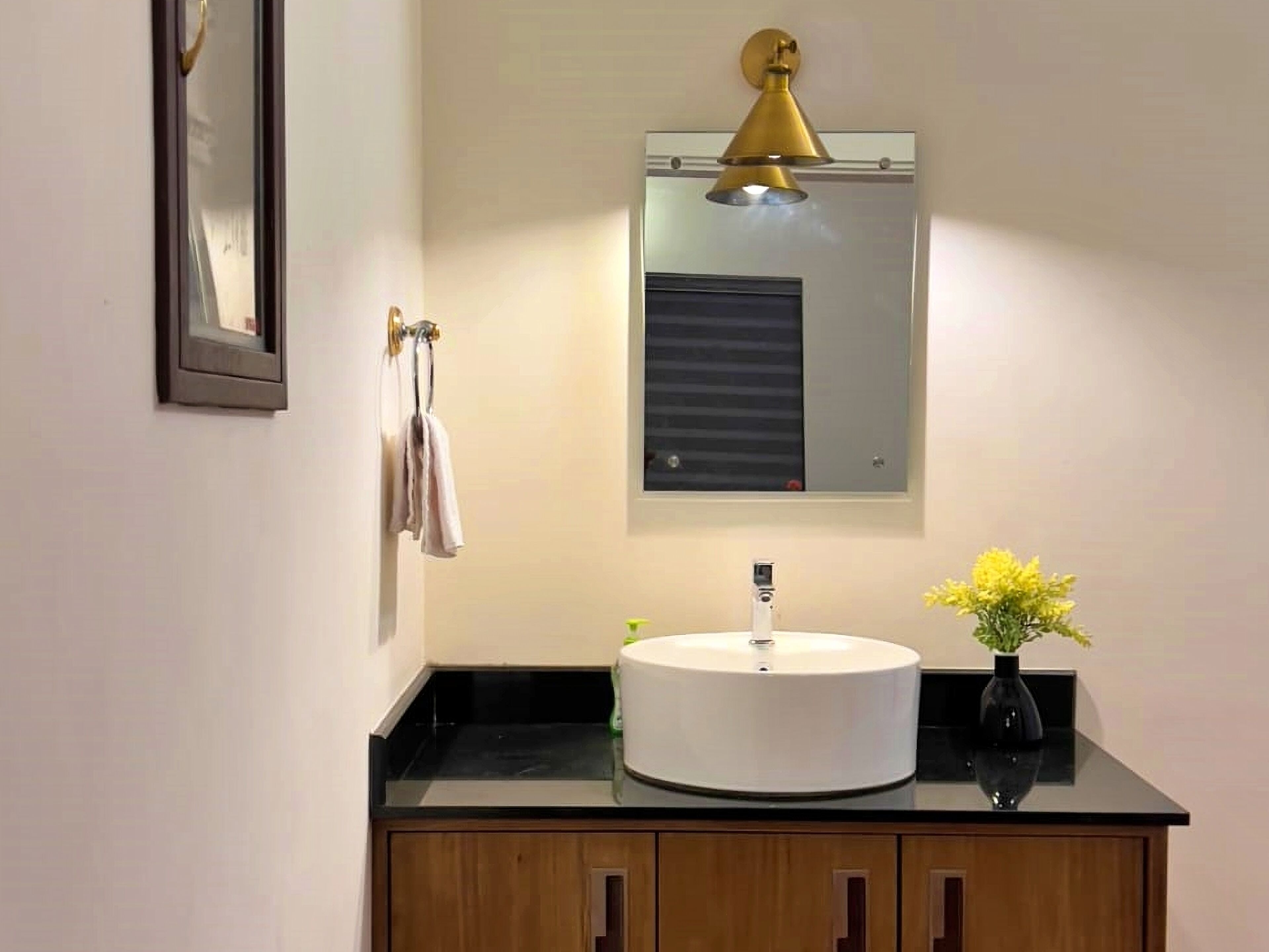Une salle de bain avec un lavabo rond blanc, un luminaire suspendu doré, un miroir et un vase rempli de fleurs jaunes.