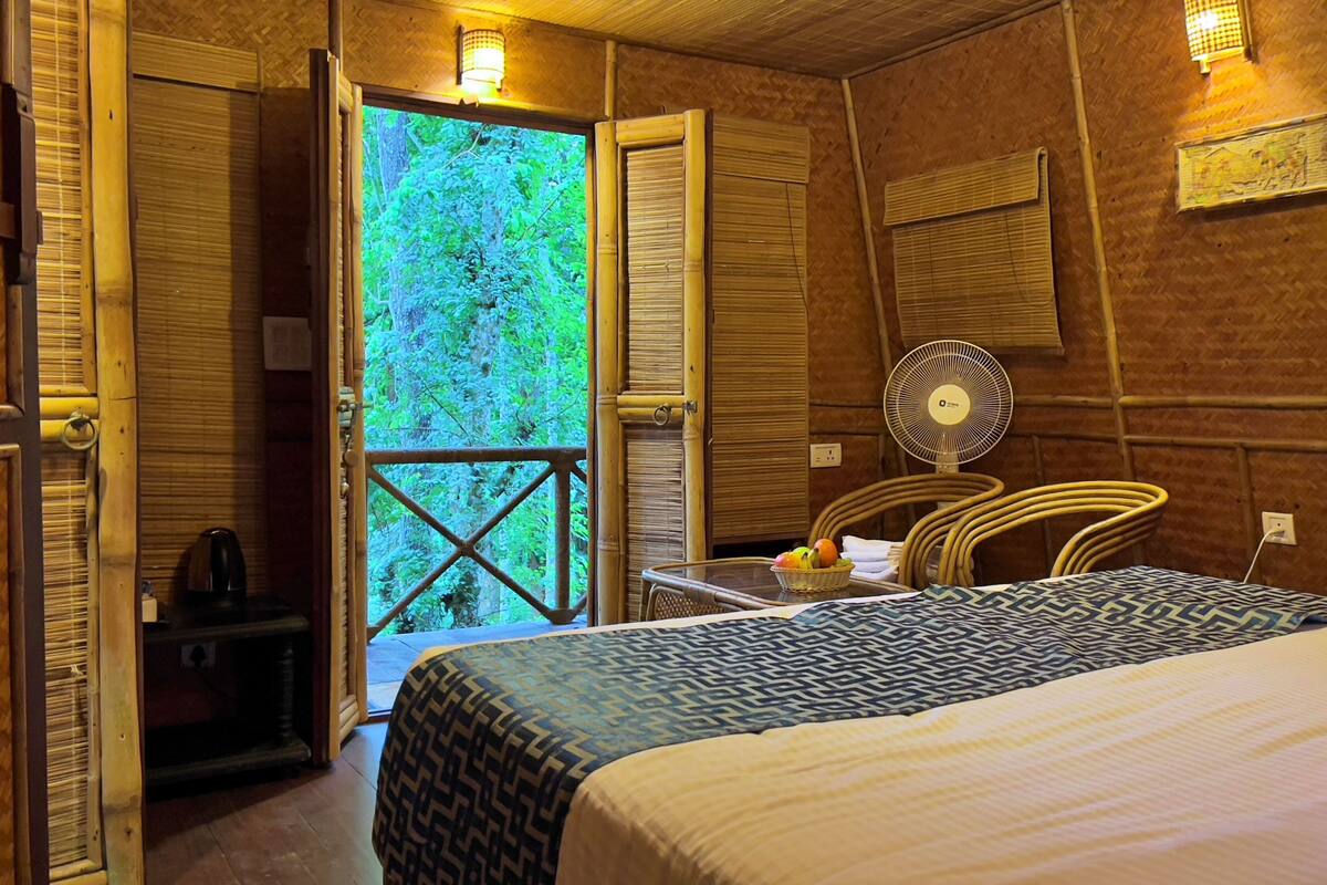 A room with a bed, a table with a basket of fruit, a fan, and a view of greenery through an open door.