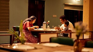 Une salle à manger avec une table dressée pour trois personnes. Un vase rempli de fleurs est posé sur la table, et la pièce dégage une atmosphère chaleureuse grâce à un luminaire fixé au mur.
