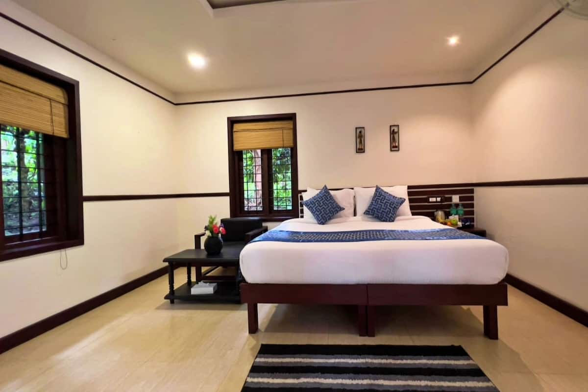 A modern bedroom with a large bed, a small table with a vase, and a window with blinds.