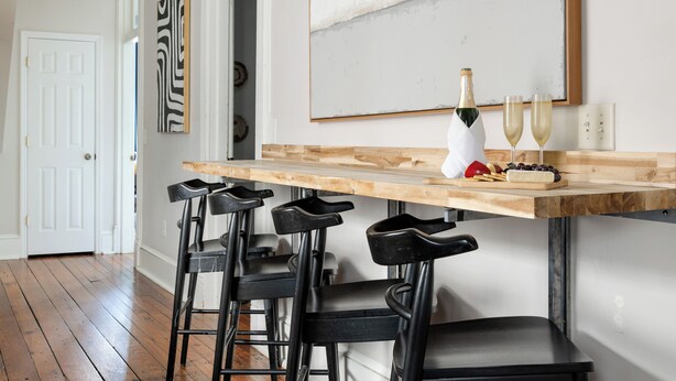 Una cocina con encimera de madera, taburetes negros tipo barra, una botella de vino y dos vasos.