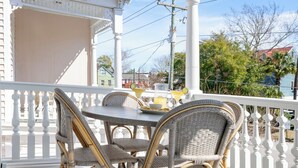 Un porche cubierto con sillas de mimbre y una mesa, barandillas blancas y vistas a una casa y árboles.