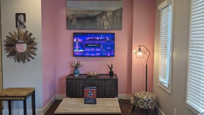 Una sala de estar con un televisor montado en la pared, una mesa consola con adornos, una lámpara de pie y un ventanal con persianas.
